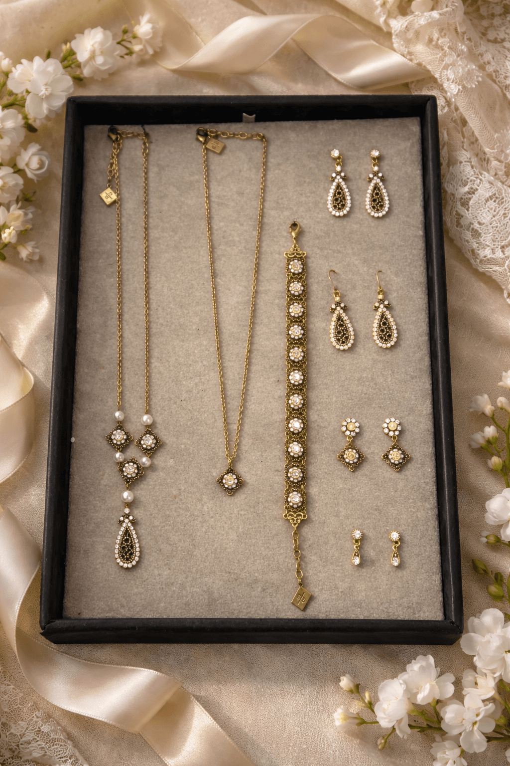 Goldener Schmuck mit Kristallen und Perlen in einem schwarzen Tablett, umgeben von weißen Blüten.