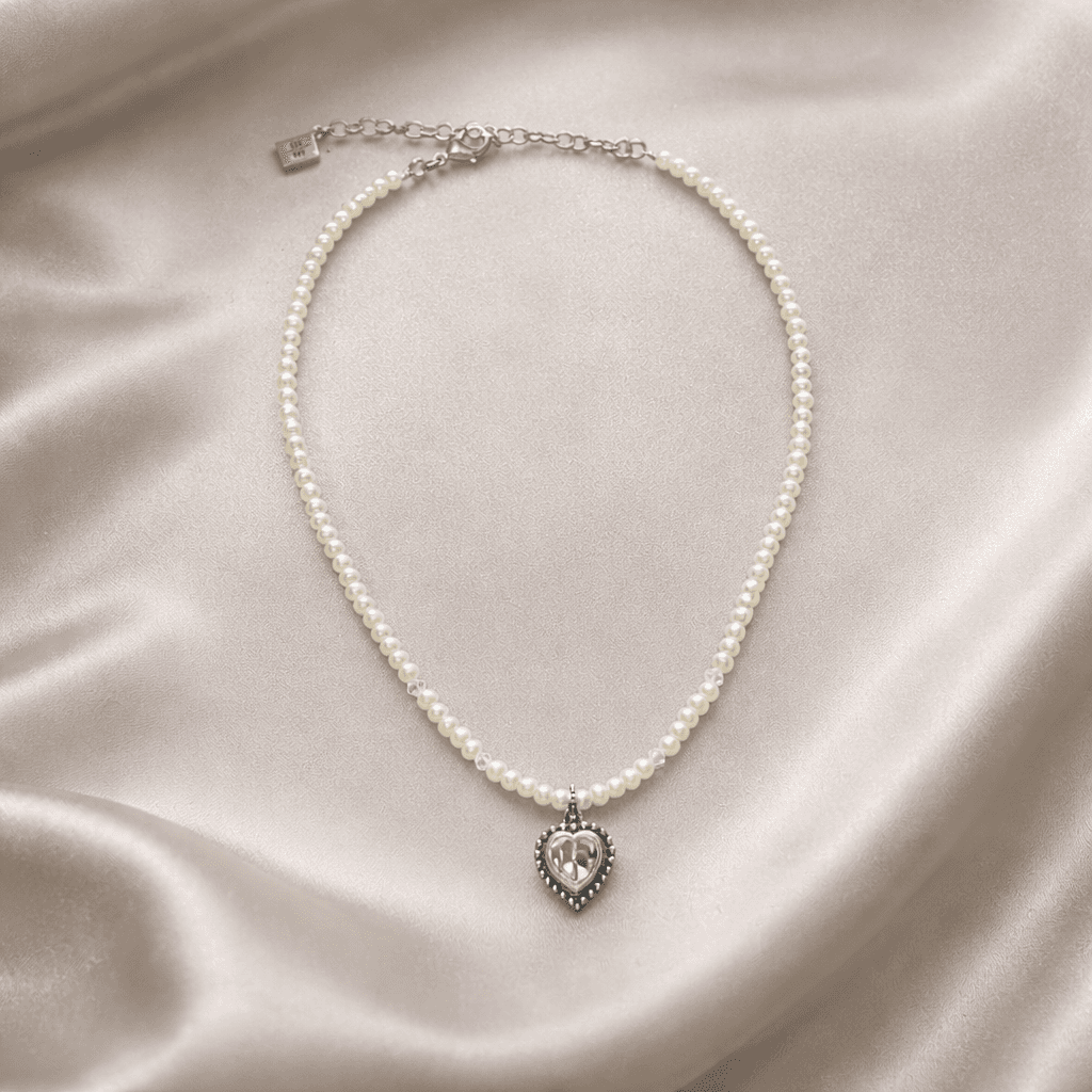 Elegante Perlenkette mit silbernem Herzanhänger auf einem sanft gewellten, cremefarbenen Satin-Hintergrund.