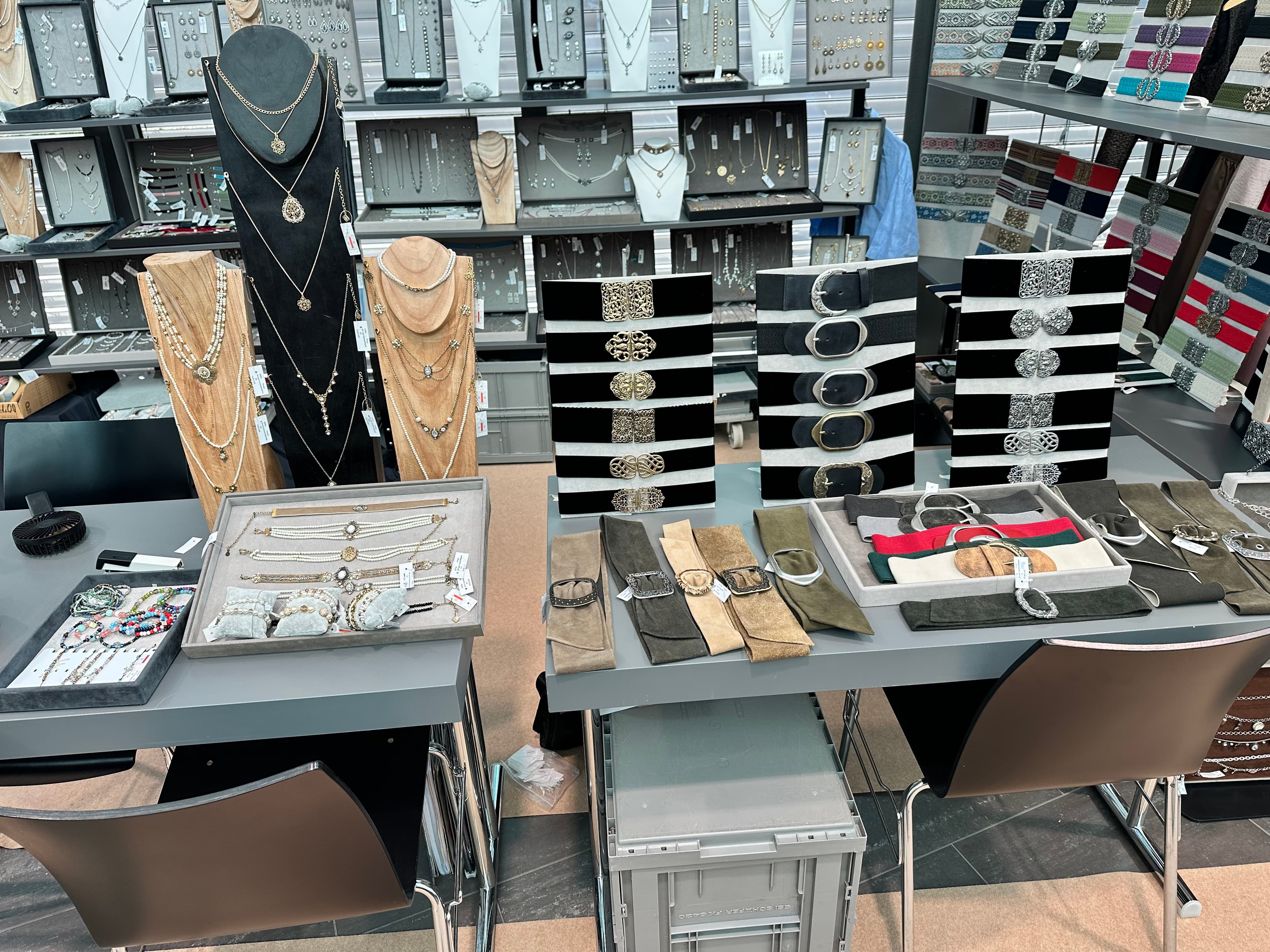 Verkaufsstand mit eleganten Halsketten auf Büsten, Gürteln auf Ständern und Schmuckstücken in grauen Tabletts.